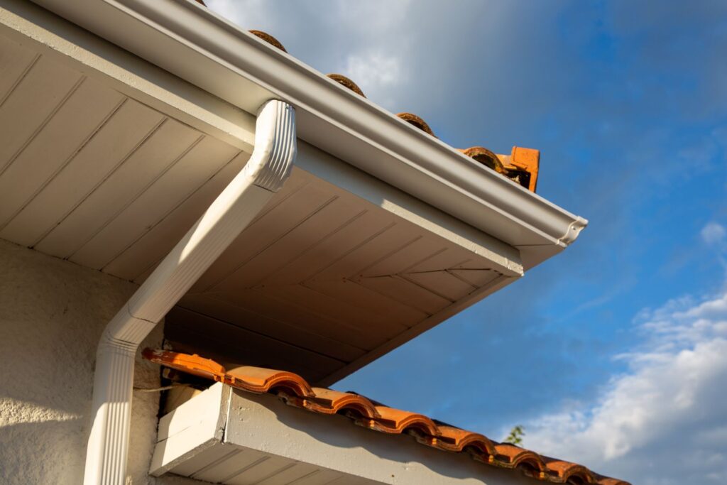 A close-up of a house corner shows white gutters and clay roof tiles against a partly cloudy sky.
