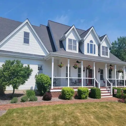 Large white house with gabled roof and front porch, surrounded by a manicured lawn and landscaping under a clear blue sky.