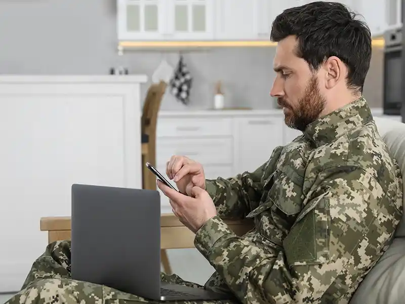 A person in military uniform uses a smartphone, sitting beside a laptop in a modern kitchen setting, appearing focused and contemplative.
