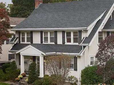 The image depicts a two-story house with a dark roof, white siding, and black shutters, surrounded by trees and shrubs.