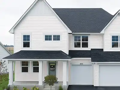 White two-story house with black roof, large windows, and twin garages. Small porch with potted plants in front. Overcast sky.