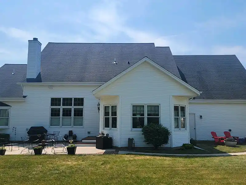 White house with dark roof, patio furniture, and green lawn. Clear sky above, quiet residential setting. No recognizable landmarks or historical buildings visible.