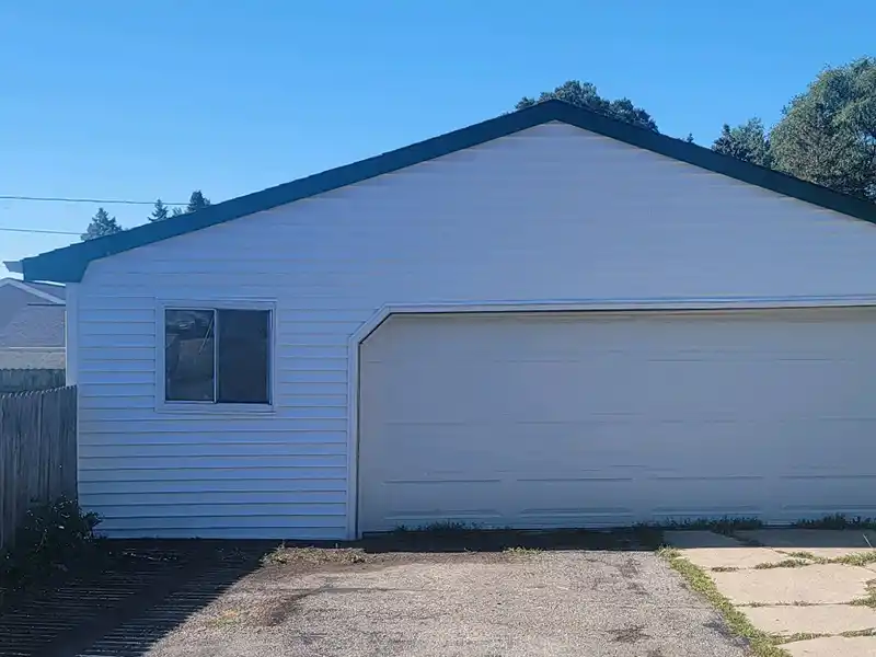 A white garage with a closed door, single window, and a wooden fence on the side under a clear blue sky.